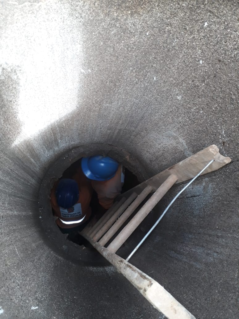 Dois trabalhadores estão no poço de uma galerias. Apenas os capacetes azuis e parte do macacão laranja são vistos de cima. Estão ao lado de uma escada, colocada para a descida. O buraco mal cabe os dois. A parede do tubo é escura, com o concreto já envelhecido. 